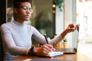 Concentrated African American woman in casual clothes sitting at wooden table with laptop while writing down information about work in planner